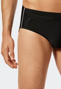 Schwarze Schwimmslips mit einem Reißverschlussfach, weißen seitlichen Akzenten, glatter Textur und einer enganliegenden Passform, die den Taillen- und Oberschenkelbereich betont.