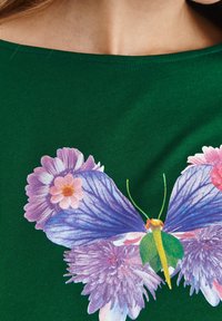 Zelená bavlněná košile s velkým grafikou motýla v purpurové a modré barvě, obklopená květinovými akcenty v růžové a purpurové.