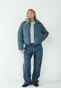 Puchowa kurtka w ciemnoszarym kolorze z wysokim kołnierzem, z pikowanymi sekcjami. Dopasowana z luźnymi niebieskimi dżinsami oraz czarnymi butami wsuwanymi.