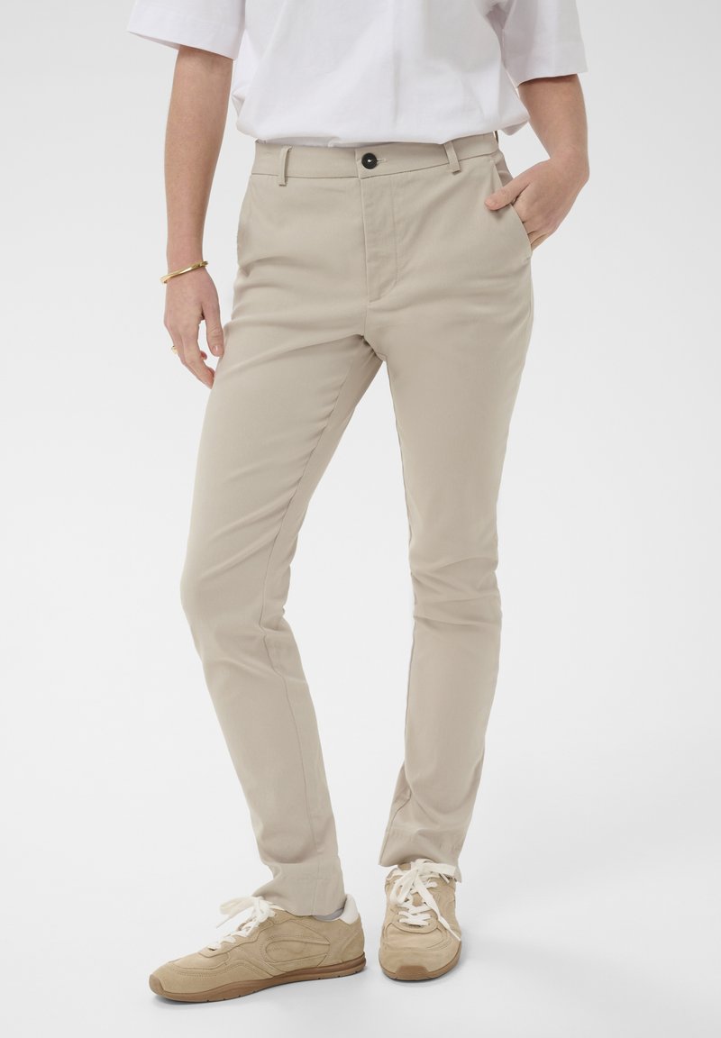Pantalons beige slim-fit avec une texture lisse, dotés d'une poche avant, d'une fermeture à bouton, portés avec des baskets beige texturées.