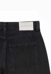Jean en denim noir montrant la poche arrière et une étiquette blanche avec des caractères japonais cousue à la ceinture.