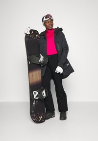 Snowboarder in dark jacket and pants, bright pink top, holding a snowboard with a dark wood and graphic design. Wearing white gloves and goggles.