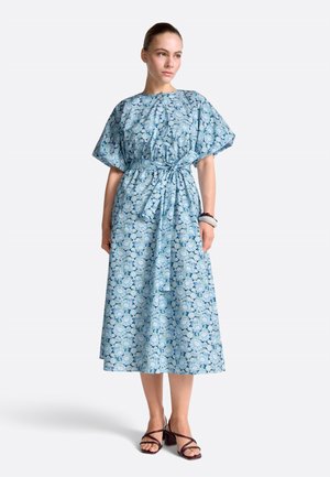 Kobieta w niebieskiej, kwiecistej sukience midi z krótkimi rękawami, zawiązaną kokardą w talii i czarnych sandałach na obcasie, stojąca na jednolitym tle.