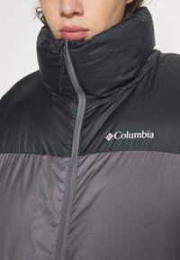 Giacca a trapunte Columbia nera e grigia. Presenta un colletto alto, chiusura con zip e tessuto texturizzato con un logo sul petto.