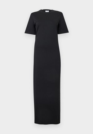 Mujer con cabello oscuro y tatuajes, vestida con un vestido maxi negro de manga corta, sosteniendo un clutch negro y llevando zapatos slingback blancos de punta puntiaguda.