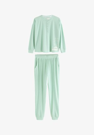 Nahaufnahme einer Person, die ein hellgrünes strukturiertes Langarmshirt und eine Hose trägt, auf der Seite mit „BY TED BAKER“ bestickt.