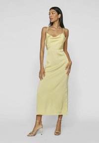 Vestido midi de satén amarillo con escote de cowl, finos tirantes y una silueta ajustada. Combinado con tacones metálicos con tiras.