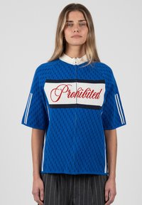 Mujer con top azul de manga corta con cremallera y la palabra "Prohibited" en escritura roja en el pecho, con patrones de rayas verticales en las mangas y los lados.