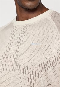 Μπεζ αθλητικό μπλουζάκι φτιαγμένο από υφασμάτινη ύφανση, με διακριτό μοτίβο από κουκίδες και γραμμές, και  με μικρό μπλε λογότυπο Nike στο στήθος.