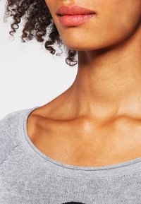 Šedý pletený top se širokým výstřihem, charakterizovaný strukturovanou tkaninou a viditelnými detaily na ramenech a klíčních kostech.