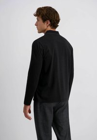 Schwarzes Langarmshirt mit geradem Schnitt und dezenten Schulternahtdetails, kombiniert mit strukturierten grauen Hosen.