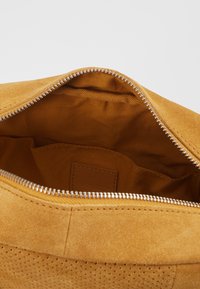 Bolsa de ante color beige con cremallera plateada parcialmente abierta, revelando un interior de tela marrón y un pequeño bolsillo interior con una etiqueta de cuero cosida.