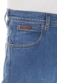 Vaqueros de mezclilla azul con cintura de altura media, que presentan una etiqueta de marca de cuero marrón, costuras naranjas y estilo de cinco bolsillos.
