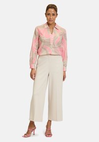 Langärmlige Bluse mit pinkem abstrakten Muster, kombiniert mit beigen, weit geschnittenen Hosen. Das Modell trägt pinke Riemchensandalen. Glatte Stofftexturen.