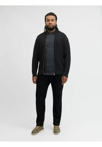 Crna rebrasta zip-up kardigana preko tamnog sive majice, uparen s crnim hlačama i zelenim cipelama sa žutim vezicama, stoji oštro protiv bijele pozadine.