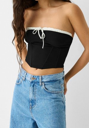 Vrouw draagt een zwarte strapless korsettop met witte rand en lichtblauwe jeans met hoge taille, staat met één hand achter haar rug.