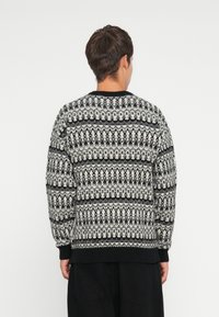 Πλεκτή ζακέτα σε γκρι και μαύρο χρώμα με πολύπλοκα γεωμετρικά σχέδια, ριγωτούς καρπούς και κλασικό σχεδιασμό γύρου λαιμού. Απαλή υφή, χαλαρή εφαρμογή.