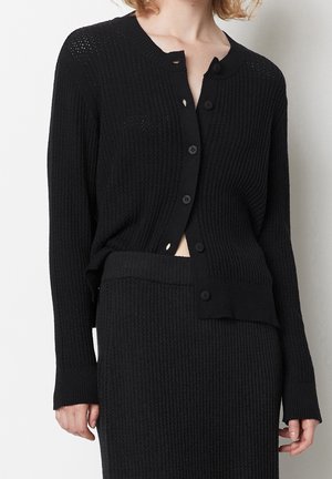 Femme portant un cardigan en tricot noir texturé à boutons et une jupe en tricot noire assortie, montrant une partie du ventre et les mains sur les côtés.