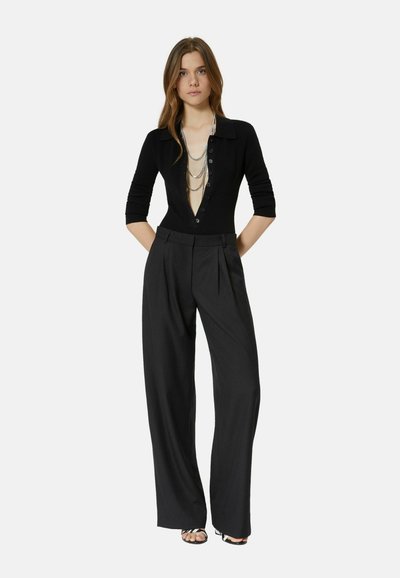 Young woman wearing black deep V-neck buttoned top, layered silver chains, wide-leg black trousers, and black strappy sandals, standing with hands behind.