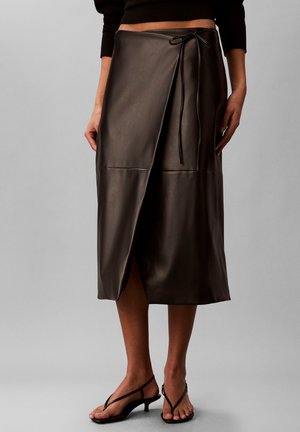 Femme portant une jupe midi portefeuille en cuir marron foncé nouée à la taille avec un nœud, associée à des sandales noires à talons bas et à lanières.