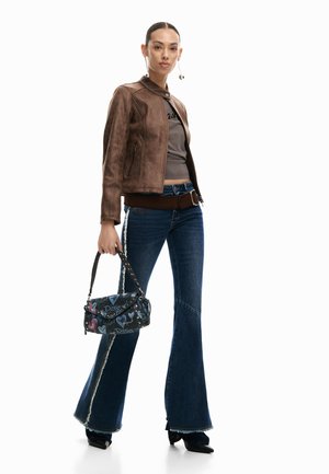 Woman wearing a brown leather jacket, grey top, dark blue flared jeans with a side stripe, black pointed shoes, holding a patterned handbag.