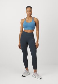 Kék sportmelltartó vékony pántokkal, magas derekú tengerészkék leggings-szel párosítva. A modellen világosszürke sportcipők vannak. Simaság jellemzi az anyagot, minimális márkajelzéssel.