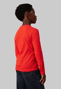 T-shirt a maniche lunghe in cotone rosso brillante, con scollo rotondo e maniche aderenti, abbinato a pantaloni in denim scuro.