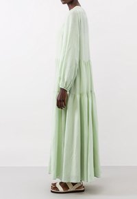 Personne à la peau foncée portant une robe longue à manches longues vert clair à volants et des sandales blanches plates à brides, debout de côté.