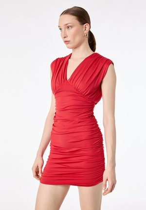 Kvinde med brunt hår i hestehale iført rød ærmeløs, rynket og figursyet kjole, gyldne øreringe i form af hoops, kigger mod venstre på hvid baggrund.