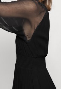 Czarna sukienka z przezroczystymi długimi rękawami, dekoltem w serek i marszczonym paskiem w talii. Fakturowana tkanina opada od talii do krawędzi.