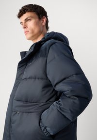 Νεαρός άνδρας με σγουρά μαλλιά που φοράει ένα ναυτικό μπλε τζάκετ πούφ με κουκούλα, στέκεται προφίλ σε λευκό φόντο.