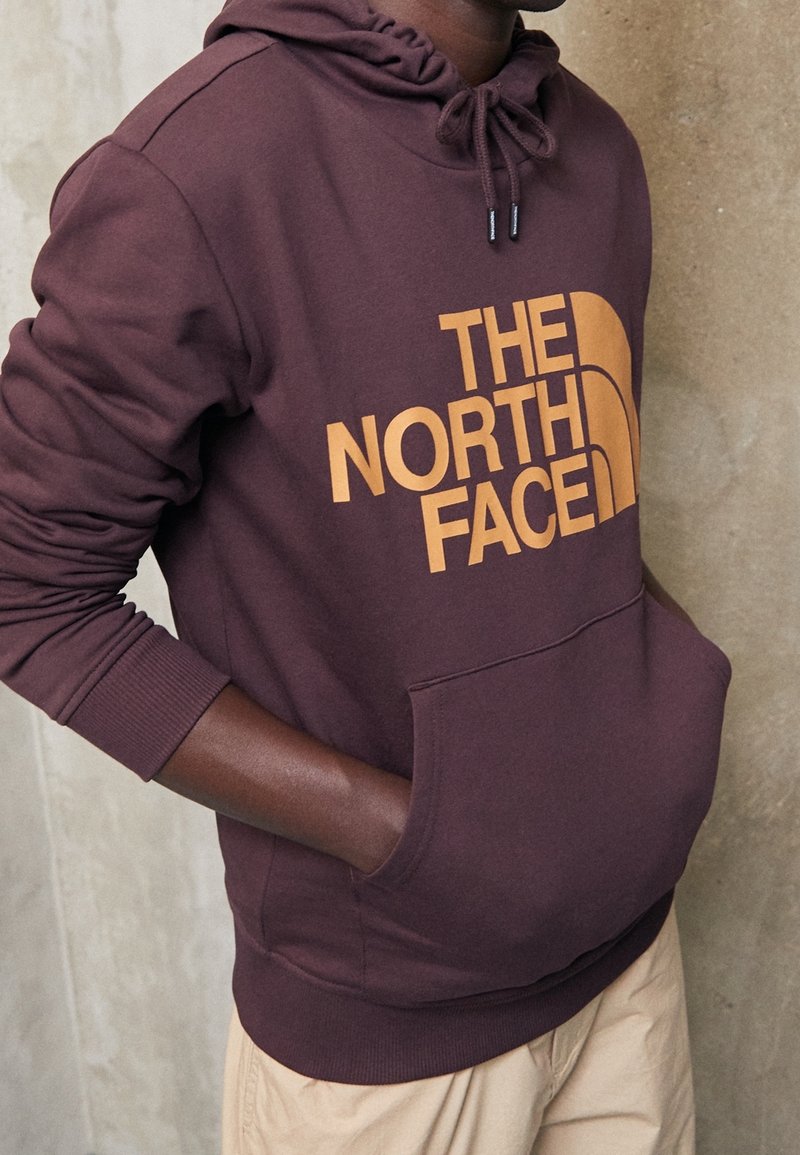 Sudadera con capucha color granate con un gran logo naranja de "THE NORTH FACE". Cuenta con capucha con cordón ajustable y bolsillo canguro frontal. Textura suave de algodón.