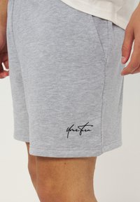 Ανοιχτό γκρι σορτς με πλαϊνές τσέπες που φοριούνται από ένα άτομο, με μικρό μαύρο κεντητό κείμενο κοντά στο τελείωμα στο αριστερό πόδι.