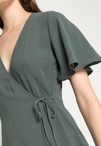 Vihreä kietaisumekko, jossa on V-aukkoreuna ja lyhyet liehuvat hihat. Valmistettu sileästä kankaasta, jossa on sidottava vyötärö, joka mahdollistaa säädettävän istuvuuden.