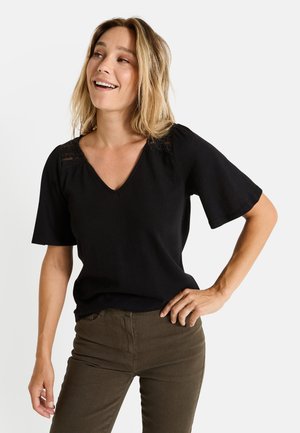 Top negro de cuello en V con mangas cortas, que presenta detalles de encaje en los hombros. Combinado con pantalones ajustados verde oliva. Textura de tela suave.