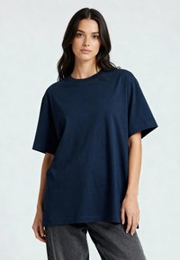 Tamsiai mėlynos spalvos medvilnės marškiniai trumpomis rankovėmis, laisvas kirpimas, apvalus iškirpimas ir lygus paviršius. Dėvimi su pilkais tekstūruotais kelnėmis.