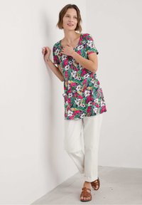 Gėlių tunika su daugiaspalviu raštu tamsiame fone, trumpomis rankovėmis ir laisvu siluetu. Suderinta su baltais kelniais ir rudais sandalais.