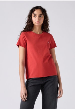 Νέα γυναίκα με σγουρά μαλλιά που φοράει ένα απλό κόκκινο κοντομάνικο t-shirt και μαύρα τζιν, στέκεται μπροστά σε ουδέτερο φόντο.