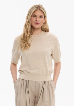 Kobieta o długich blond włosach, ubrana w beżowy krótki sweter z krótkim rękawem i luźne, plisowane beżowe spodnie, z rękami w kieszeniach.