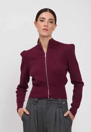Frau mit zurückgebundenen dunklen Haaren, die einen bordeauxroten Pullover mit Reißverschluss und graue plissierte Hose trägt, die Hände in den Taschen.