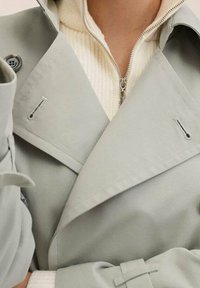 Jasnoszary płaszcz trencz z wyraźnym kołnierzem, dwurzędowym krojem i guzikami. Nałożony na kremowy sweter z zamkiem błyskawicznym o prążkowanej strukturze.