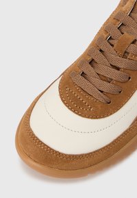 Ruskeaa mokkanahkaa oleva päällinen, jossa on kermaisia nahkaisia yksityiskohtia, pyöreät nauhat ja kuvioitu kumipohja; reunojen kohdalla ompelut.