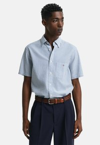 Kurzärmliges, knöpfbares Hemd mit blau-weißen Streifen, aus Baumwollstoff, mit einer linken Brusttasche, getragen mit dunkelblauen Hosen und einem braunen Gürtel.