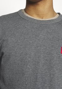 Detailní záběr na osobu v šedém svetru s kulatým výstřihem, který má přes béžovou košili, viditelný je krk a horní část hrudníku.