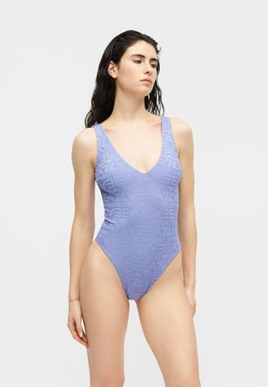 Młoda kobieta ubrana w jasnofioletowy jednoczęściowy kostium kąpielowy z nadrukiem marki, stojąca na tle jednolitego jasnego tła.