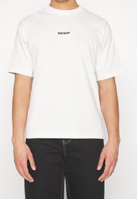 T-shirt en coton blanc à manches courtes, col rond, présentant un logo texte "WRSTBHVR" noir sur la poitrine, coupe décontractée et texture lisse.