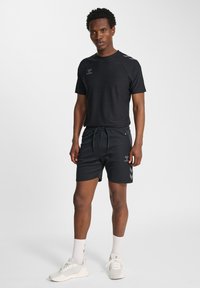 Schwarzes sportliches Kurzarmshirt und Shorts aus leichtem Stoff, mit Logo und Chevron-Akzenten, kombiniert mit weißen Turnschuhen und Socken.