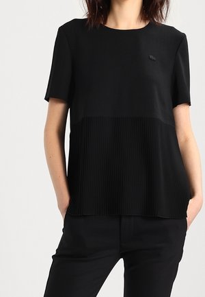 Vrouw draagt een zwart topje met korte mouwen met een geplooid onderste deel en bijpassende zwarte broek, handen in de zakken, staand tegen een witte achtergrond.