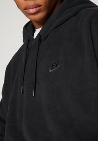 Črna flis kapa s kenguru žepom, kapuco s snemnimi vrvicami in vezenim logotipom Nike na prsih. Mehka tekstura in sproščen kroj.