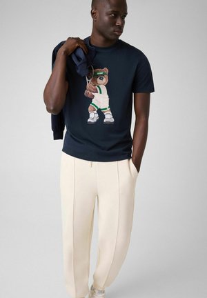 Uomo che indossa una t-shirt blu navy con un orsacchiotto in tenuta da basket, tiene una giacca blu navy sulla spalla e pantaloni color crema con le mani nelle tasche.